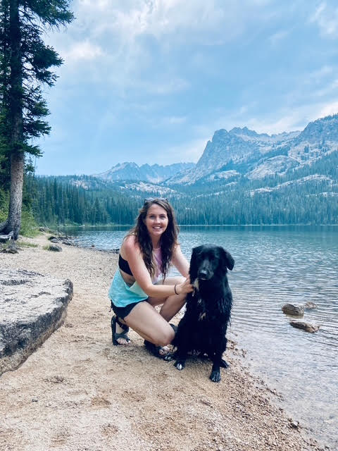 A woman kneels beside a black dog on a sandy lakeshore with mountains and pine trees in the background.