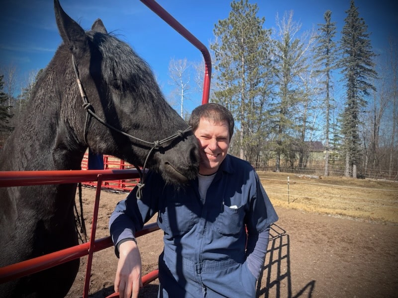 A man in blue coveralls smiles while standing next to a black horse by a red metal fence, with trees and a blue sky in the background.