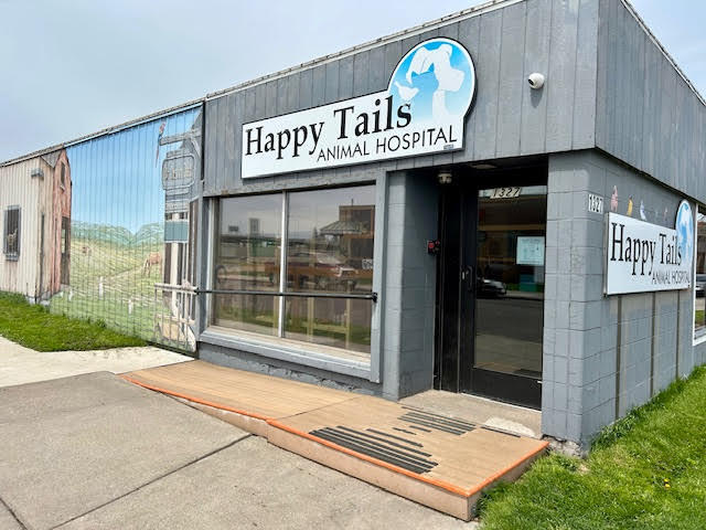 Gray building with signs reading "Happy Tails Animal Hospital." Large front window, ramp access, and a mural of a barn scene on the side wall.