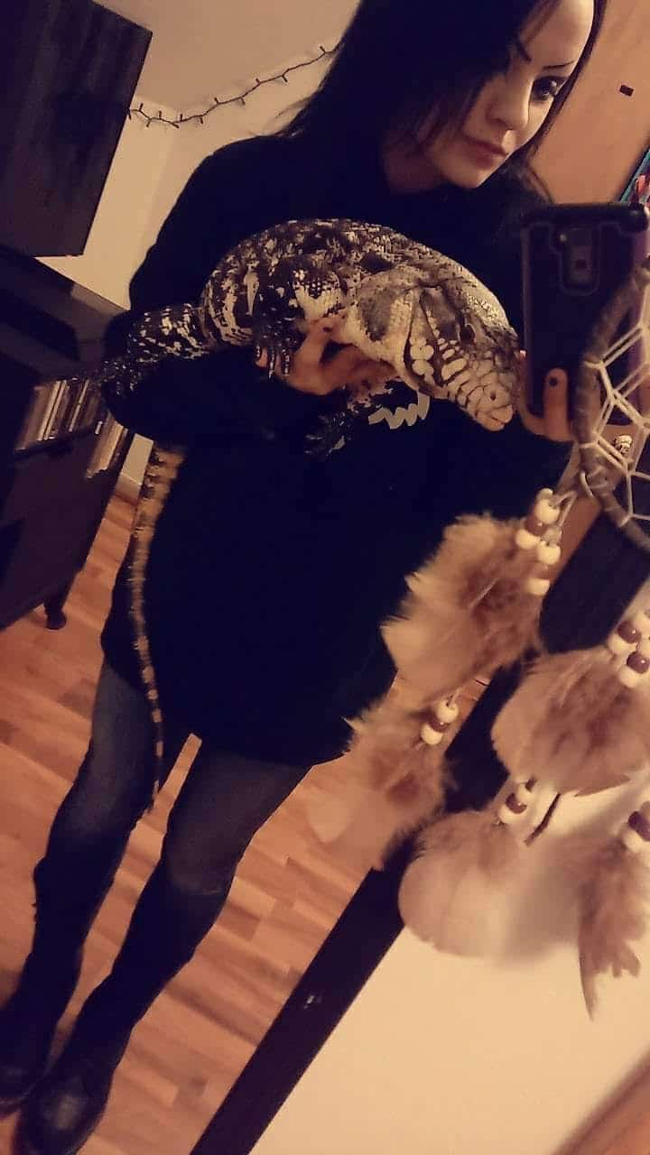 A person taking a mirror selfie holds a large lizard, possibly a tegu, in one arm. A dreamcatcher and string lights are visible in the background.