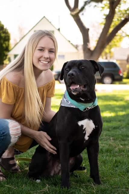 A young woman with long blonde hair kneels on grass next to a black dog wearing a green bandana. They are both outdoors in a sunlit yard.